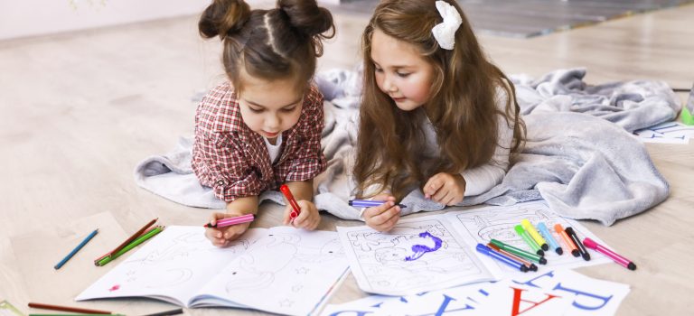 Two tiny cute girls which are drawing in the coloring book laying on the floor on the blanket and learning letters